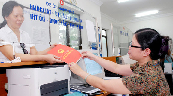 Tại văn phòng nhà nước, một cán bộ nữ mặc áo trắng đang đưa sổ đỏ cho một phụ nữ trung niên.