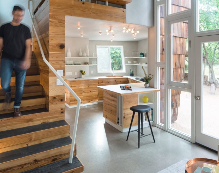 Khu bếp nhỏ bố trí khéo léo dưới tầng 1. Khu bếp với các mảng tường và tủ ốp gỗ, bên trái là cầu thang có bóng một người đàn ông đang bước xuống.