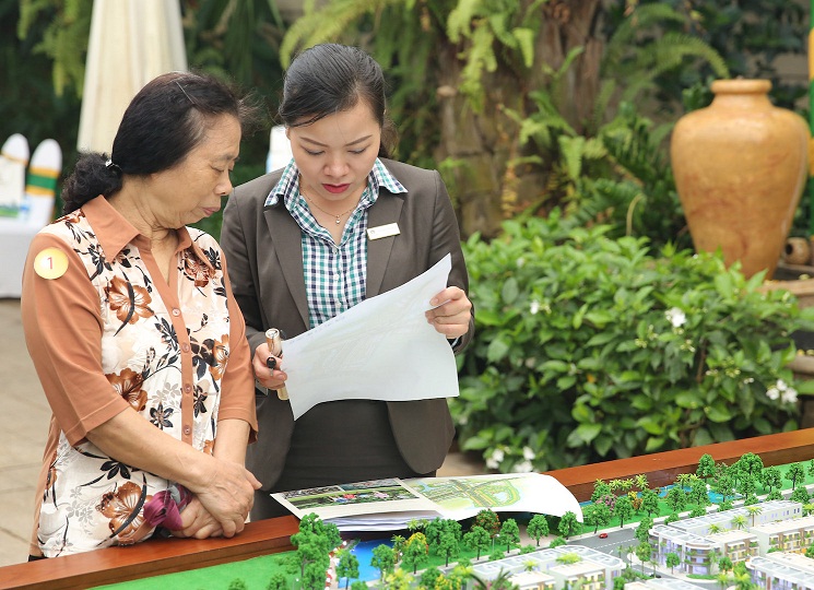 Hai phụ nữ đứng cạnh nhau xem chung một tờ giấy, cạnh sa bàn dự án bất động sản.